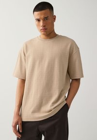 Young man with short hair wears beige textured short-sleeve shirt and black pants, standing with one hand in pocket against white background.