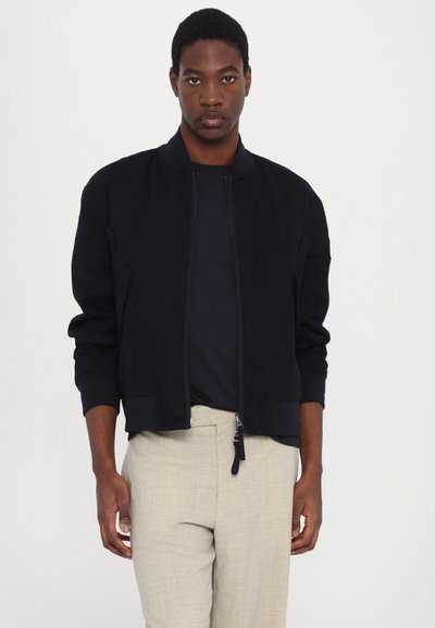 Marineblau bomberjacke mit Reißverschluss, gerippten Bündchen und Seitentaschen, kombiniert mit beigen Hosen für einen kontrastierenden Look.