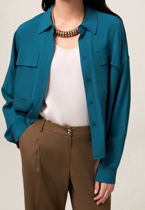 Femme portant une chemise bleu sarcelle boutonnée sur un haut blanc, un pantalon marron et un collier épais en or, debout avec une main dans la poche.