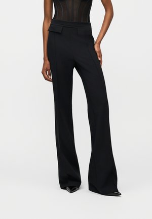 Žena oblečená v čiernych širokých nohaviciach s vysokým pásom a detailmi s klapkovými vreckami, v kombinácii s priesvitným čiernym priliehavým topom a čiernymi špicatými topánkami.