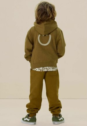Olijfgroene hoodie met een groot rond ontwerp op de achterkant, gecombineerd met bijpassende broek en een gestreept shirt, afgemaakt met grijze sneakers.