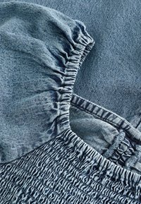 Denimkjole med en samlet ruffleærme, smocked bodice og en lys blå vask. Inkluderer strukturerede detaljer og kontrasterende syninger.