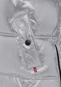Glanzende zilveren pufferjack met een zwart drukknop-flapzak en een klein rood label met een wit dierenlogo.