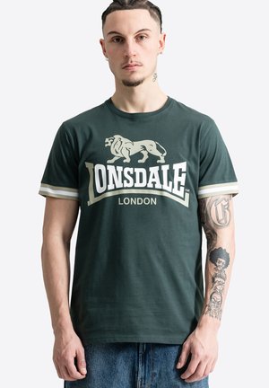 Homme aux cheveux tressés portant un t-shirt vert foncé Lonsdale London, montrant une manche tatouée détaillée sur le bras droit.