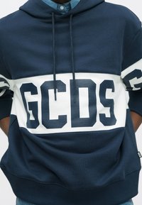 Personne portant un sweat à capuche bleu marine et blanc avec « GCDS » en grandes lettres bleu marine sur la poitrine et les manches.