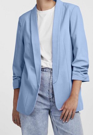 Person wearing a light blue blazer with rolled sleeves over a white t-shirt and high-waisted light wash jeans, standing against a plain background.