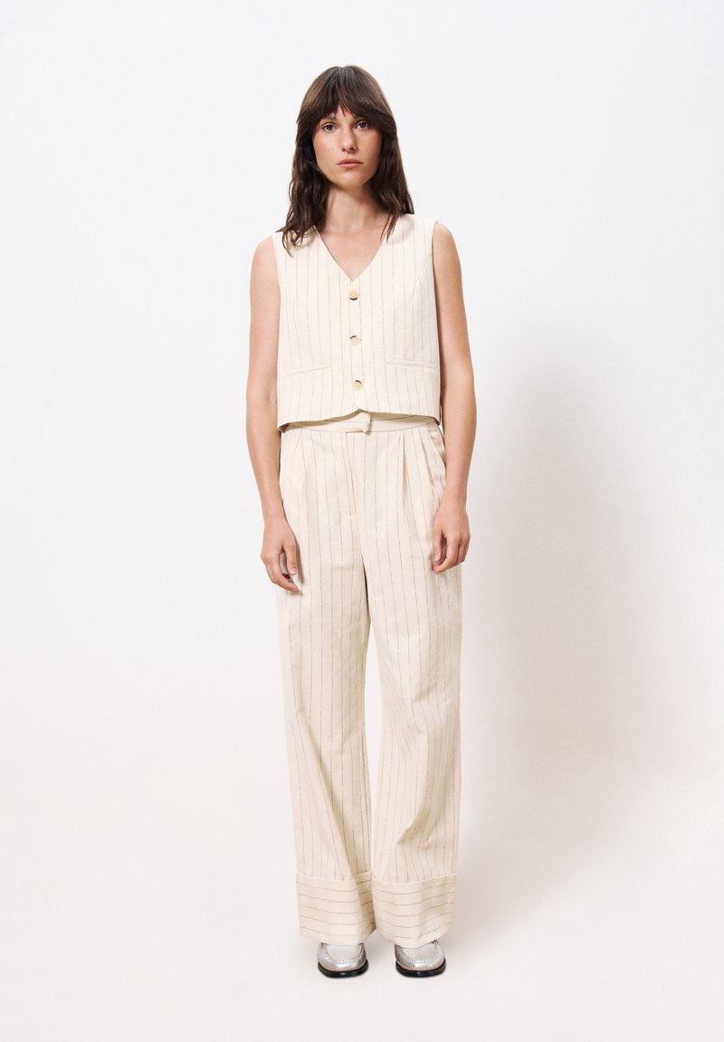 Crème-kleurige stoffen tweedelige outfit met verticale strepen; mouwloos vest met kraag en wijde broek, aangevuld met glanzende zilveren schoenen.