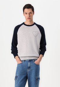 Gray and navy sweater with raglan sleeves, a ribbed hem, and a stitched heart detail on the chest, paired with light blue jeans.