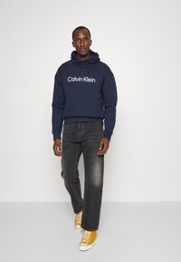 Navy hooded sweatshirt with white "Calvin Klein" logo, paired with dark gray jeans and yellow sneakers with white soles, walking against a light backdrop.