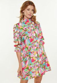 Bunt gemustertes Kleid mit hohem Kragen, Rüschensäumen und einer Mischung aus rosa, grün, blau und gelben Formen sowie floralen Designs.