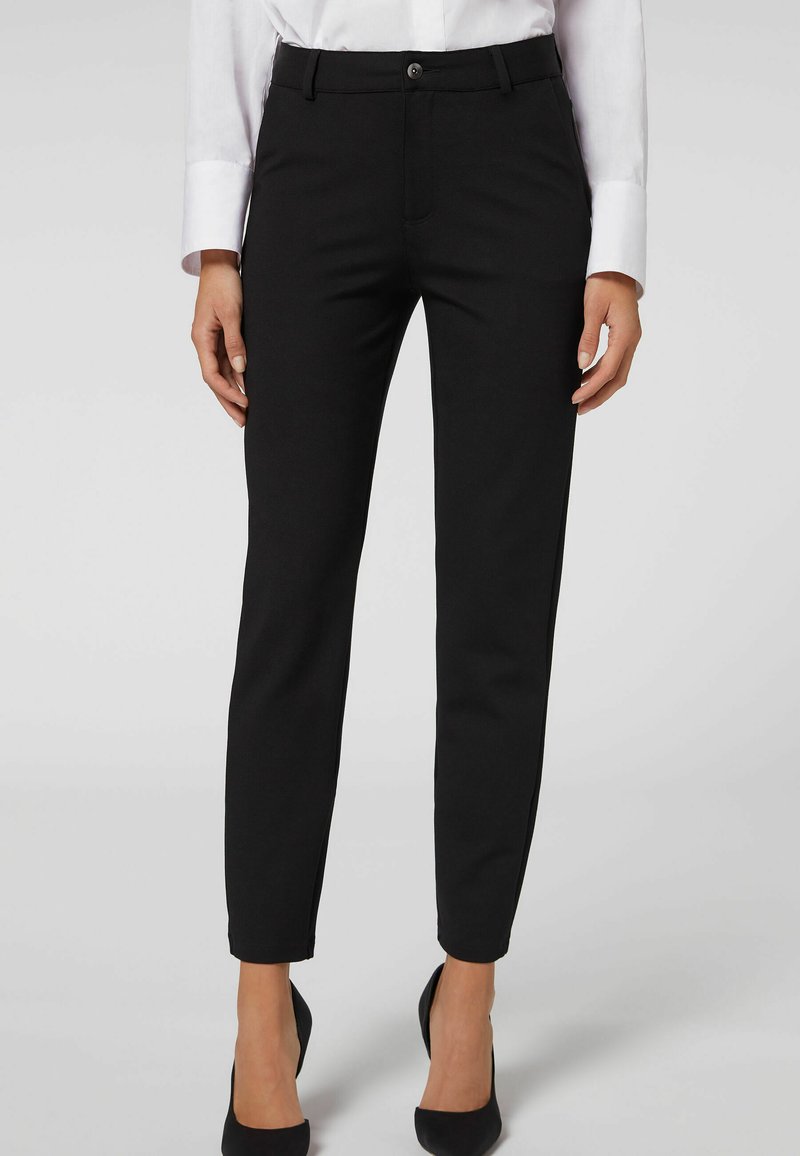Pantalones negros ajustados con una textura suave, que cuentan con un cierre de botón, trabillas para el cinturón y piernas estrechas, combinados con tacones altos negros.