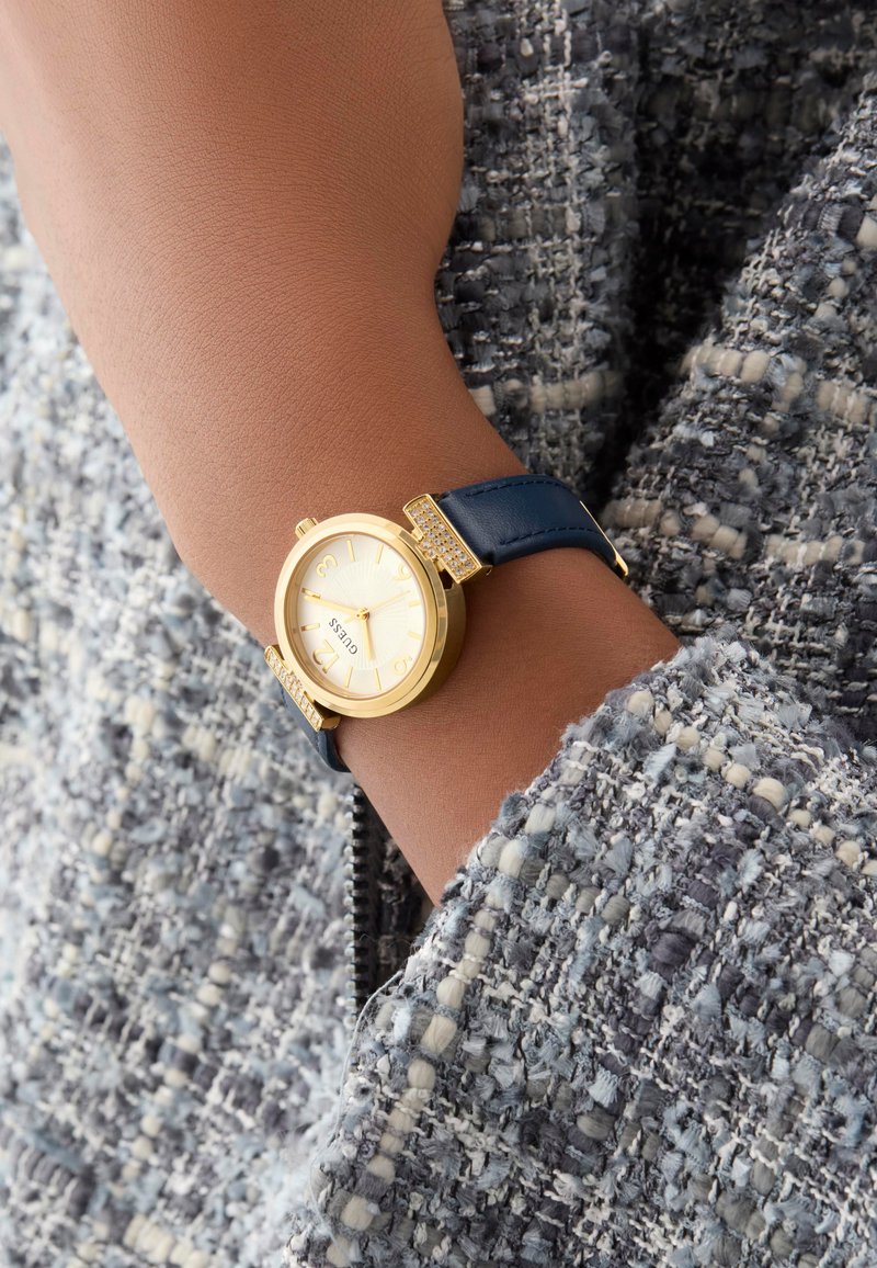Gold-tone watch with a round face, white textured dial, and blue leather strap featuring decorative accents near the lugs.