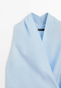 Haut en lin bleu clair sans manches avec un décolleté en V profond et un détail devant superposé sur fond blanc.