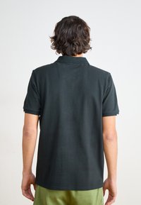 Mørkegrøn polo med klassisk krave og korte ærmer, med en ensartet tekstur og standard pasform, set fra ryggen.