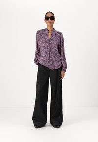 Blusa floreale viola con chiusura a bottoni e colletto alto, abbinata a pantaloni neri larghi. Il modello indossa occhiali da sole scuri, in piedi su uno sfondo neutro.