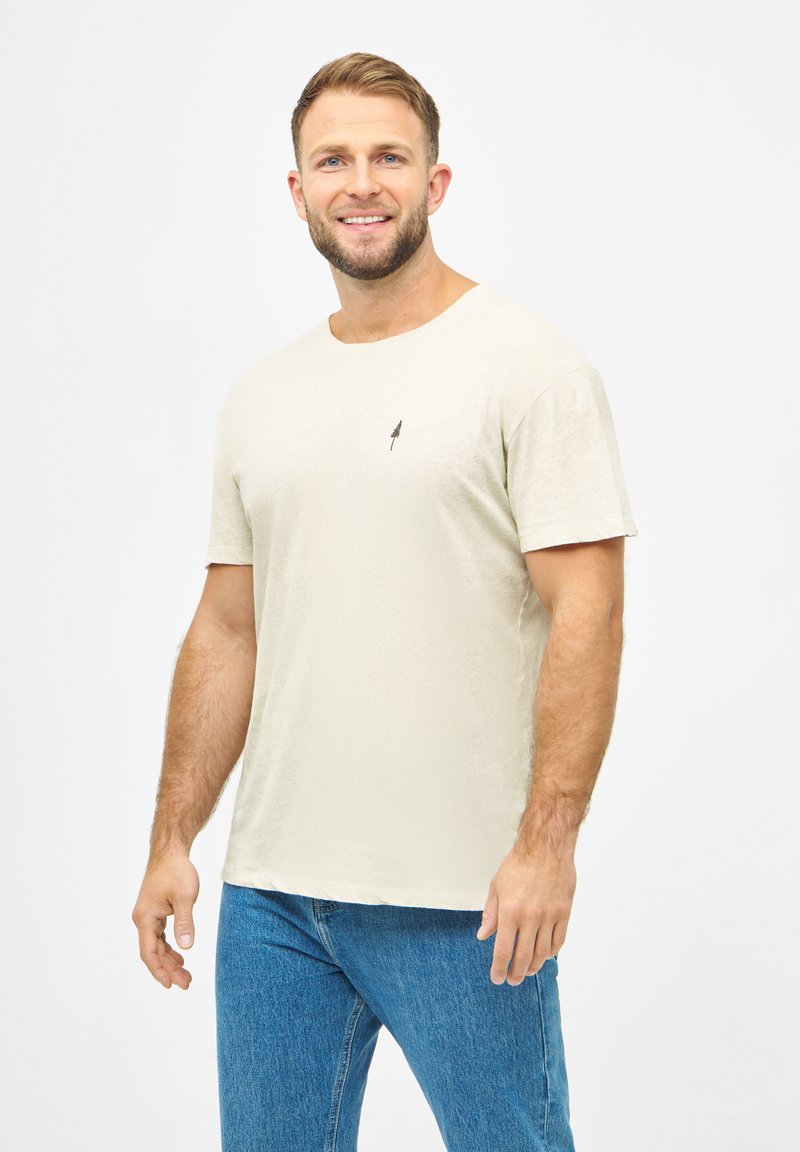 Beige T-Shirt mit kurzen Ärmeln, runder Ausschnitt und kleinem schwarzen Baum-Logo auf der Brust, getragen mit blauen Jeans.