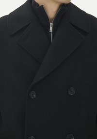 Cappotto in lana nera doppiopetto con ampio colletto, strato interno con zip in contrasto e bottoni neri rotondi. Texture liscia su tutta la superficie.