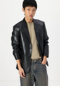 Black faux leather blazer with a notched collar, single button closure, and front pockets, paired with a light beige shirt and denim jeans.