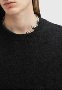 Prim-plan al unei persoane care poartă un pulover negru din tricot cu un guler destrămat și un lănțișor gros de argint.