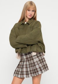 Giacca oversize verde oliva con collo e dettagli a toppa, abbinata a una gonna plissettata a quadri nei toni marrone e crema.