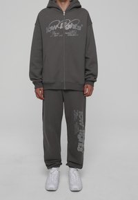 Sudadera gris con cremallera y gran logo, combinada con pantalones de chándal grises a juego con impresión de texto. Ambos de tejido suave, complementados con zapatillas blancas.