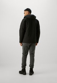 Giacca nera con cappuccio e finitura opaca, abbinata a pantaloni grigi slim. Il modello indossa sneakers nere con texture. Design semplice e funzionale.