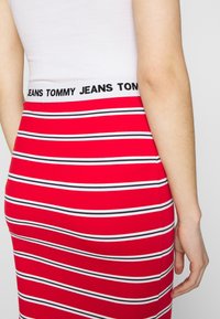 Rot gestreifter Rock in taillierter Form mit weißen und schwarzen horizontalen Streifen. Der weiße Bund trägt in schwarzer Schrift "JEANS TOMMY".