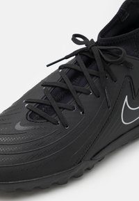 Zapatilla deportiva negra con parte superior sintética texturizada, diseño elegante, cordones finos y un logo blanco prominente en el lateral.