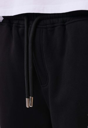 Pantaloni sportivi neri con vita elasticizzata e lacci neri con estremità metalliche, indossati con una camicia bianca parzialmente visibile sopra.