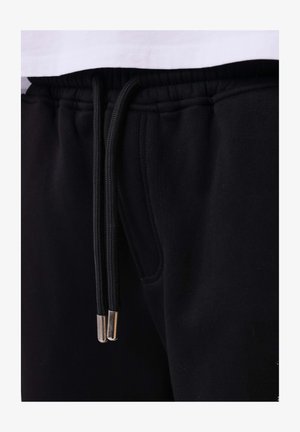 Pantalons de survêtement noirs avec une taille élastique et des cordons noirs dotés de pointes métalliques, portés avec une chemise blanche partiellement visible au-dessus.