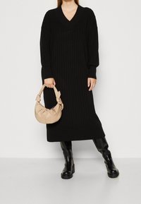 Robe noire en maille côtelée avec un col en V et des manches longues, associée à un sac à main beige avec une poignée nouée. Des bottes noires montantes jusqu'aux genoux complètent la tenue.