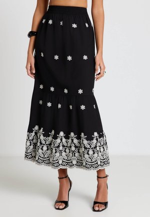 Gonna midi nera con ricamo floreale bianco sparso e denso sull'orlo, indossata con tacchi neri con cinturino alla caviglia su sfondo chiaro.