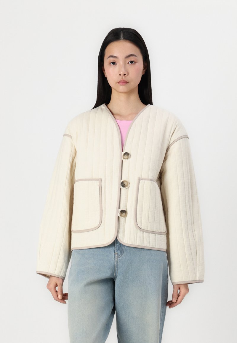 Quiltet kremfarget jakke med V-hals, tre knapper og beige detaljer. Har to store lommer foran og vide ermer. Brukt over en rosa topp.