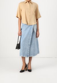 Chemise beige à manches courtes avec boutons et poche ; jupe portefeuille en denim bleu clair ; chaussures noires à talons hauts ; petit sac à main noir.