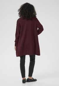 Cardigan tricoté bordeaux avec une texture en vagues, design oversize et poignets côtelés, porté par-dessus un pantalon slim noir et des mocassins noirs.