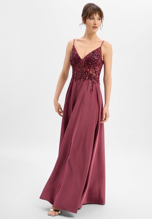 Frau in einem bodenlangen burgunderroten Kleid mit besticktem Spitzenoberteil und dünnen Trägern, die seitlich blickt vor einem schlichten Hintergrund steht.