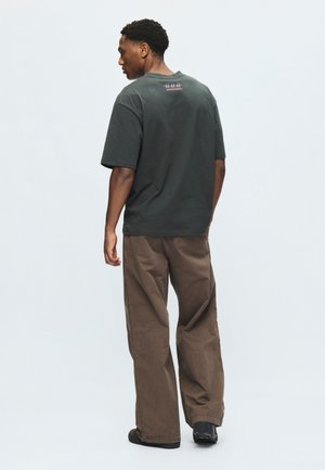 Homme de dos portant un t-shirt noir oversize avec un texte rose et un pantalon marron ample sur un fond clair uni.