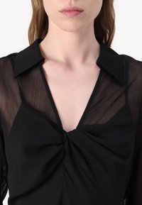 Svart blus med djup V-ringning och knuten frontdetalj, med genomskinlig texturerad tyg och spetsig krage.