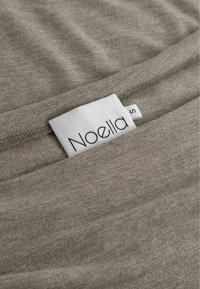 Pelēks audums tuvplānā ar mīkstu tekstūru, kura malā ir balts zīmola uzlīme "Noella" un izmērs "S".