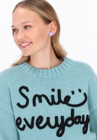 Junge Frau, die lächelt, trägt einen hellblauen gestrickten Pullover mit dem Aufdruck "Smile everyday" und lavendelfarbenen Ohrsteckern vor einem weißen Hintergrund.