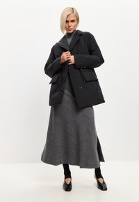 Chaqueta acolchada negra con un amplio cuello, bolsillos frontales, llevada sobre una larga falda gris. El look se completa con unos zapatos negros con recortes.