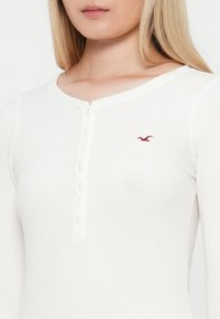 Biely pruhovaný dlhý rukáv top s okrúhlym výstrihom, detailmi s gombíkmi pozdĺž prednej strany a malým červeným logom na hrudi.