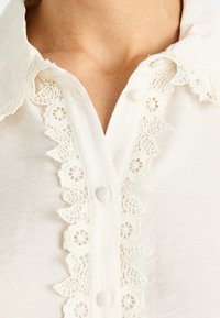 Blusa blanca de tejido trenzado con cuello redondo, delicados bordes de encaje a lo largo de la tapeta y botones redondos en la parte delantera. Textura suave.