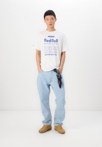 Tricou alb cu text albastru, blugi ușor bleu și teniși maro. O bandană în buzunar adaugă un detaliu contrastant de negru.