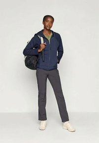 Casaco zip-up azul-marinho com capuz, camisa verde por baixo, calças cinzentas e sapatos bege. Mochila preta com alças ajustáveis.