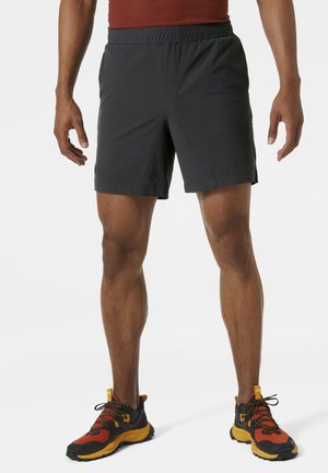 Man die zwarte sportshorts en oranje-zwart hardloopschoenen draagt, staand met iets gespreide benen op een witte achtergrond.