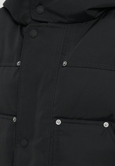 Giacca nera imbottita con bottoni a pressione, due tasche frontali con accessori in acciaio inox e una finitura opaca. Il tessuto sembra liscio e resistente.