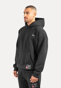 Felpa nera con cappuccio e tasca frontale, realizzata in tessuto morbido. Presenta un piccolo logo e un'etichetta sul lato inferiore sinistro.