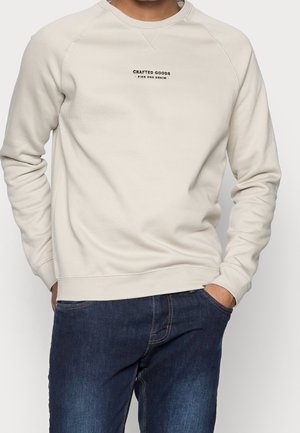 Mężczyzna w kremowej bluzie z napisem „Crafted Goods Pier One Denim” i ciemnoniebieskich jeansach, ręce w kieszeniach.