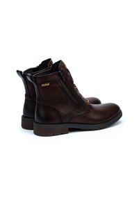 Bottines en cuir marron avec une texture lisse, fermeture à lacets et à glissière, et une semelle légèrement surélevée. Présente des accents de design minimalistes.